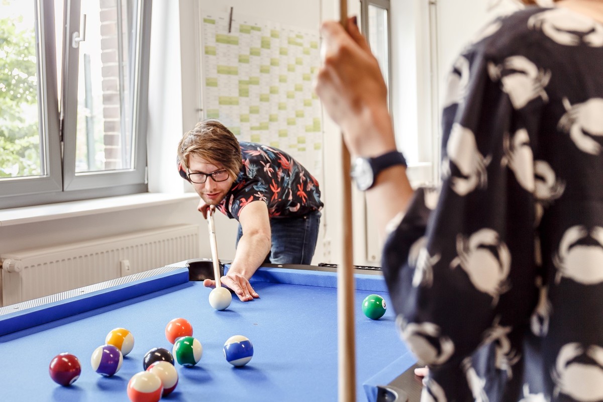 Zwei Personen spielen an einem blauen Billardtisch in einem hellen Raum.