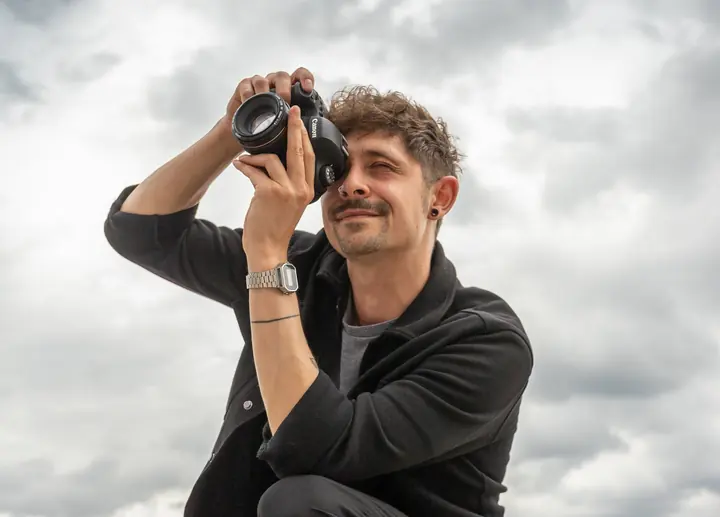 Ein Mann hält eine Fotokamera vor sein Gesicht; hinter ihm ist ein bewölkter Himmel zu sehen.