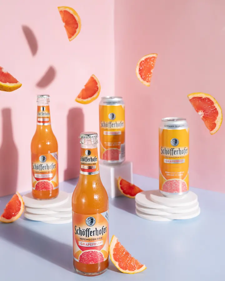 Getränkeflaschen und Dosen auf weißen Podesten mit Grapefruitstücken vor rosa Hintergrund.
