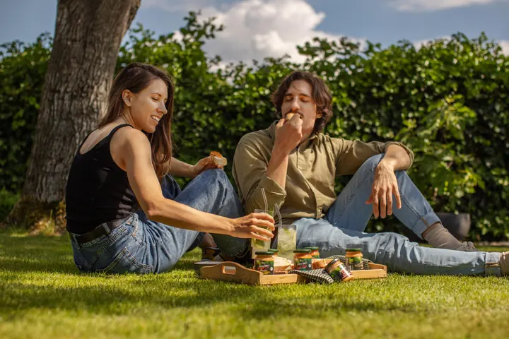 Zwei Personen sitzen mit Brot, Aufstrichen und Getränken bei einem Picknick unter einem Baum.