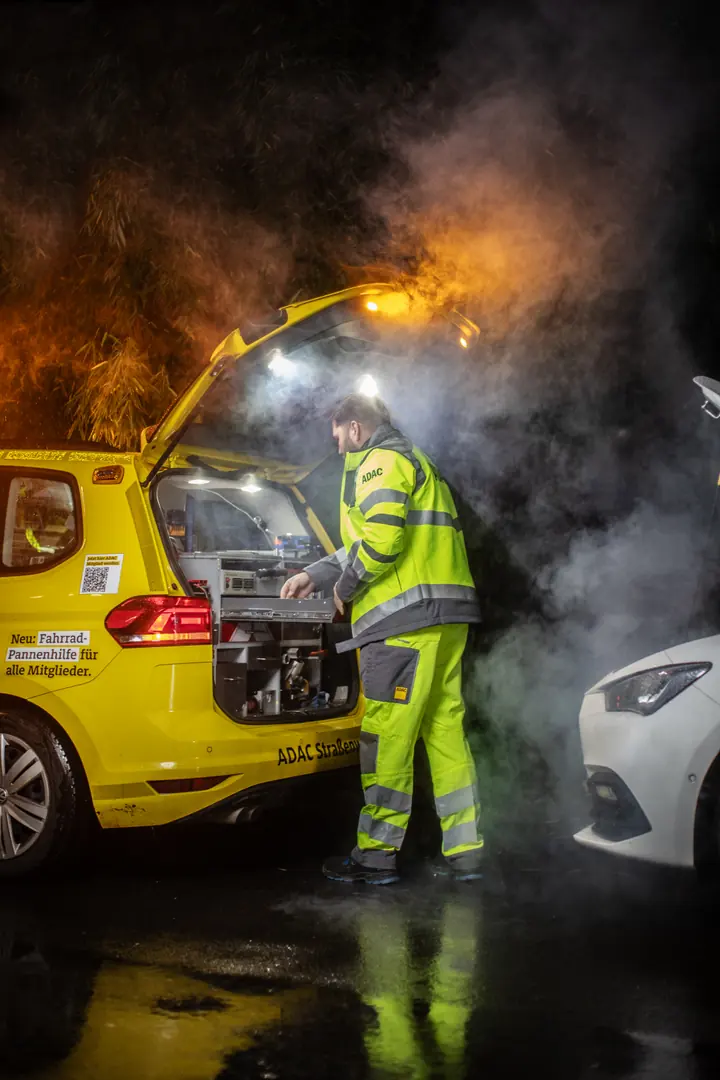 Eine Person in gelber Warnkleidung steht bei Nacht am geöffneten Heck eines gelben Servicefahrzeugs, umgeben von Licht und Nebel.