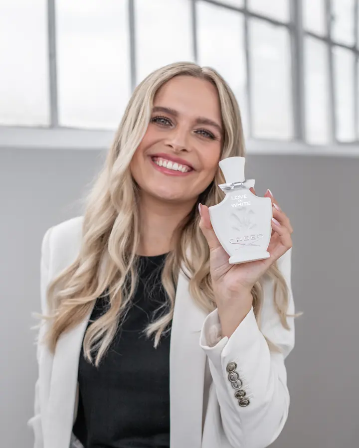 Eine lächelnde Frau in weißem Blazer hält einen weißen Parfumflakon neben ihr Gesicht.