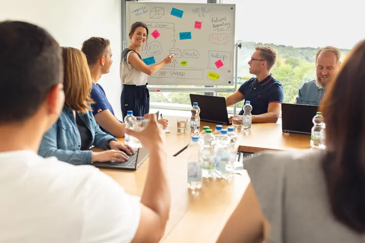 Eine Frau erläutert Notizen am Whiteboard, während mehrere Personen an einem Tisch mit Laptops sitzen.